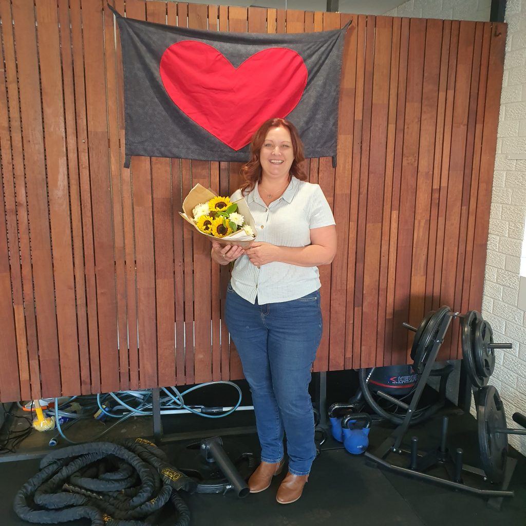 Kate Cairnduff PT wearing blue jeans and a green shirt infront of a brown wooden wall underneath a love lismore red heart with a bunch of flowers she has recieved and gym equipment on the floor