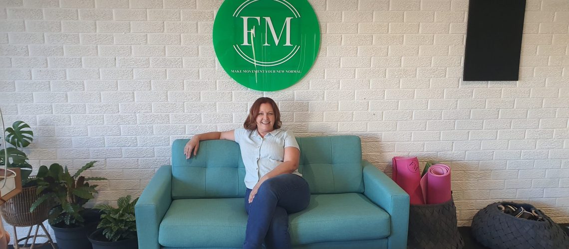Kate Cairnduff a lady personal trainer sitting on a green couch underneath the logo for FM on a white wall